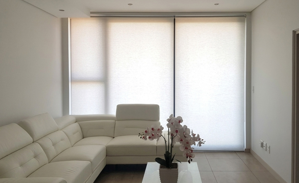 A wide interior view of automated roller blinds with light filtering fabric installed in a modern Johannesburg home with a clean cream leather sectional couch.