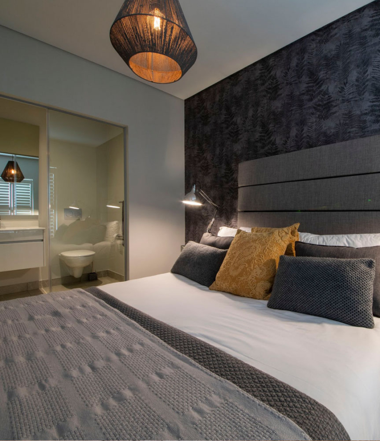 Custom monochromatic bedroom design at Serengeti Estate featuring dark botanical wallpaper, mirrored pedestal, and mustard accent cushions.