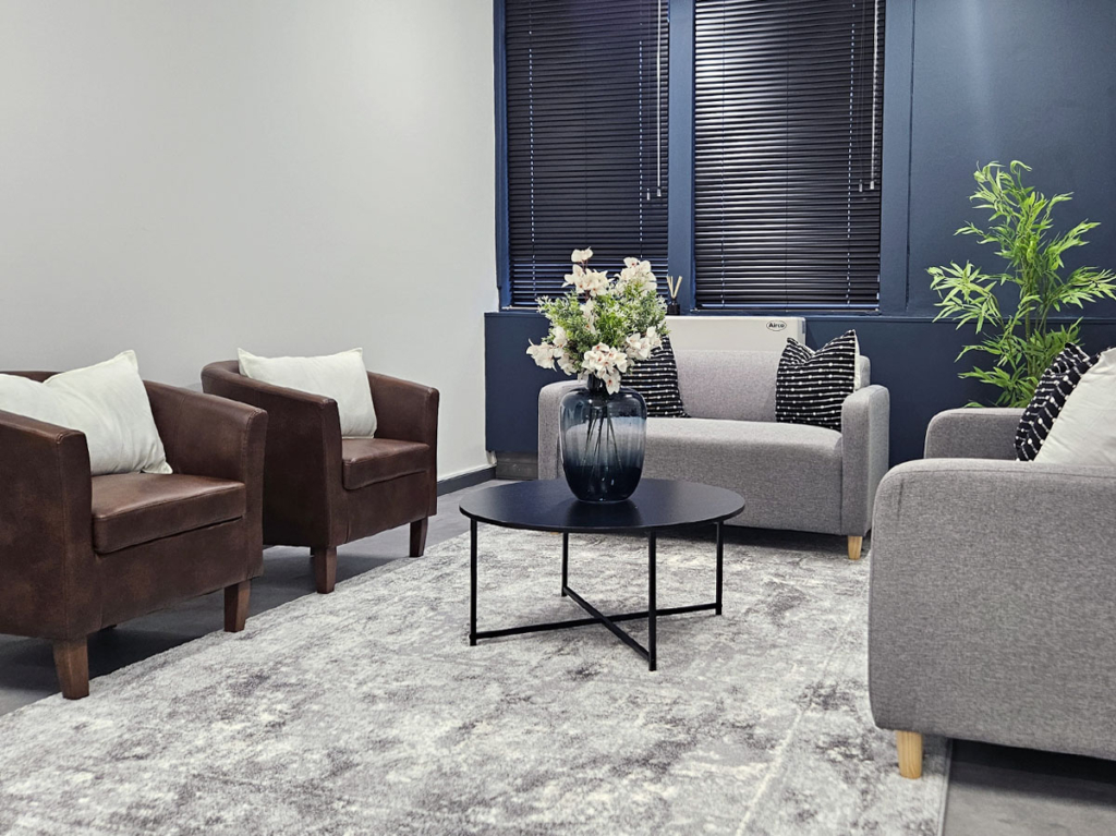 Medical consultation room lounge featuring brown leather armchairs, grey fabric couches, a black circular coffee table, and navy blue window wall with blinds.