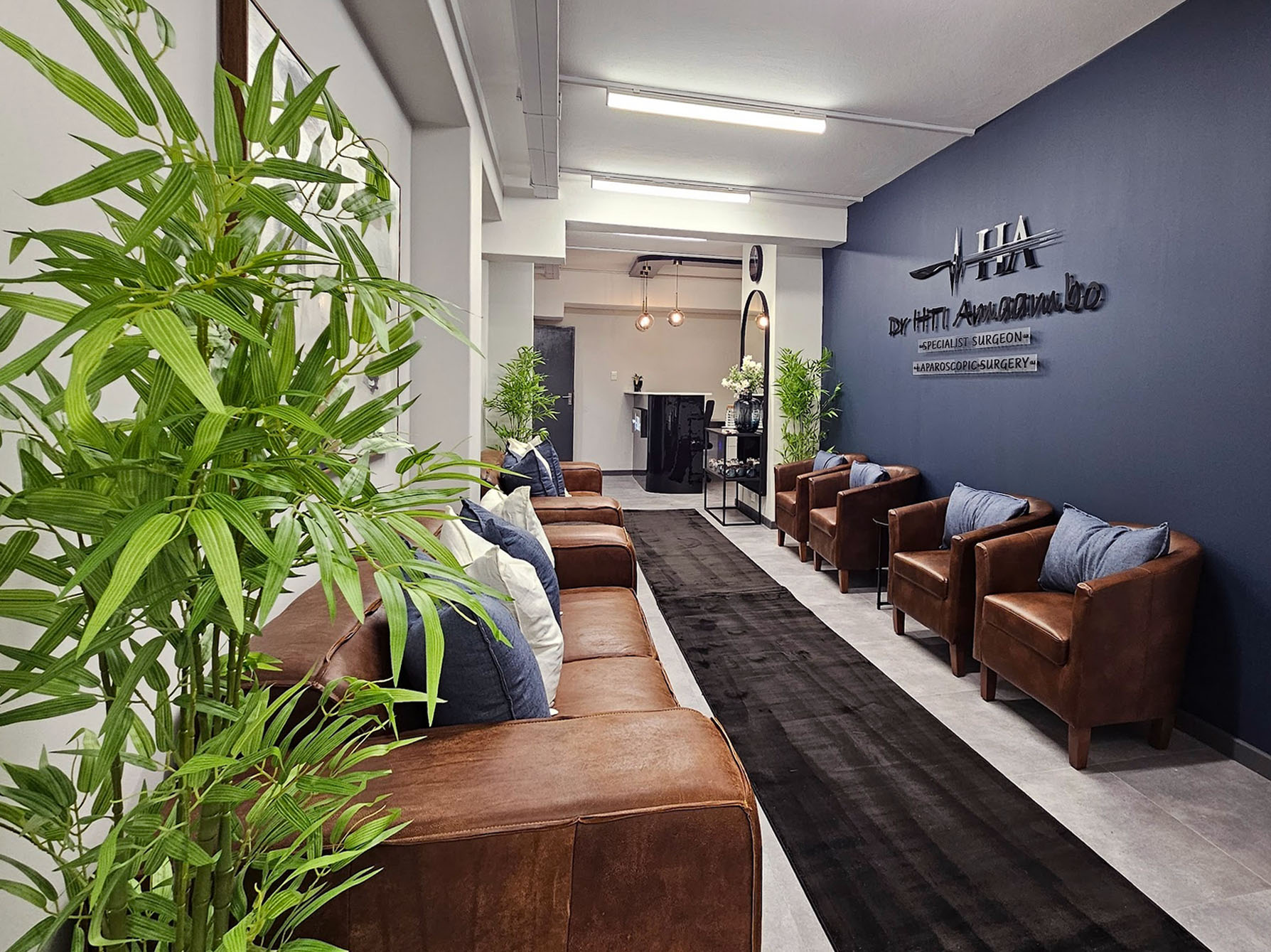 Long view of a medical waiting room featuring brown leather seating, a navy blue feature wall with 3D surgical branding, and indoor greenery.
