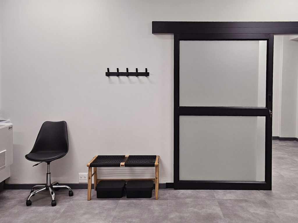Industrial-style black sliding barn door with frosted glass in a medical suite, featuring a minimalist bench and black office chair.