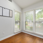 Bright corner office or studio space featuring white horizontal blinds, light grey walls, and warm timber flooring.