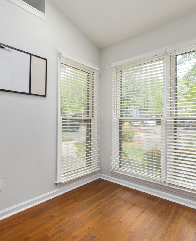 Bright corner office or studio space featuring white horizontal blinds, light grey walls, and warm timber flooring.