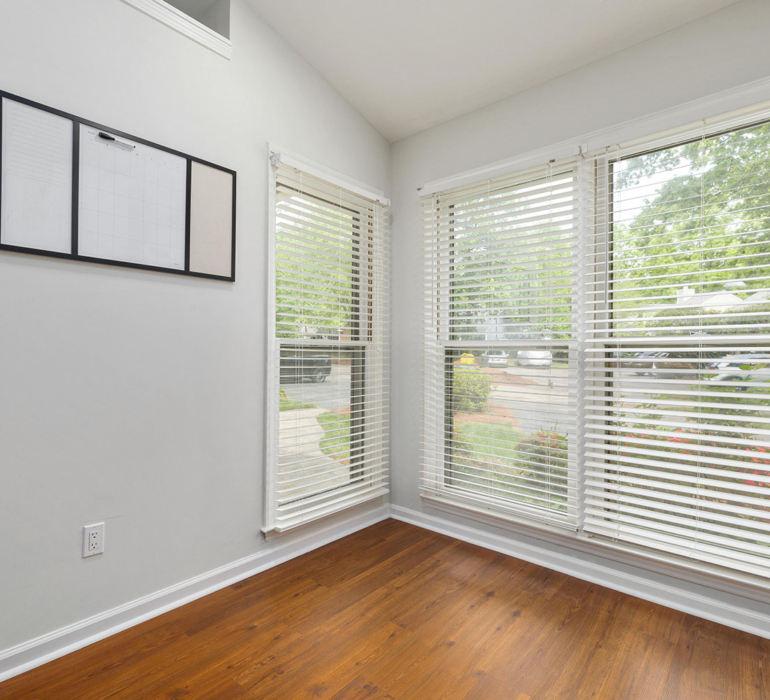 Bright corner office or studio space featuring white horizontal blinds, light grey walls, and warm timber flooring.