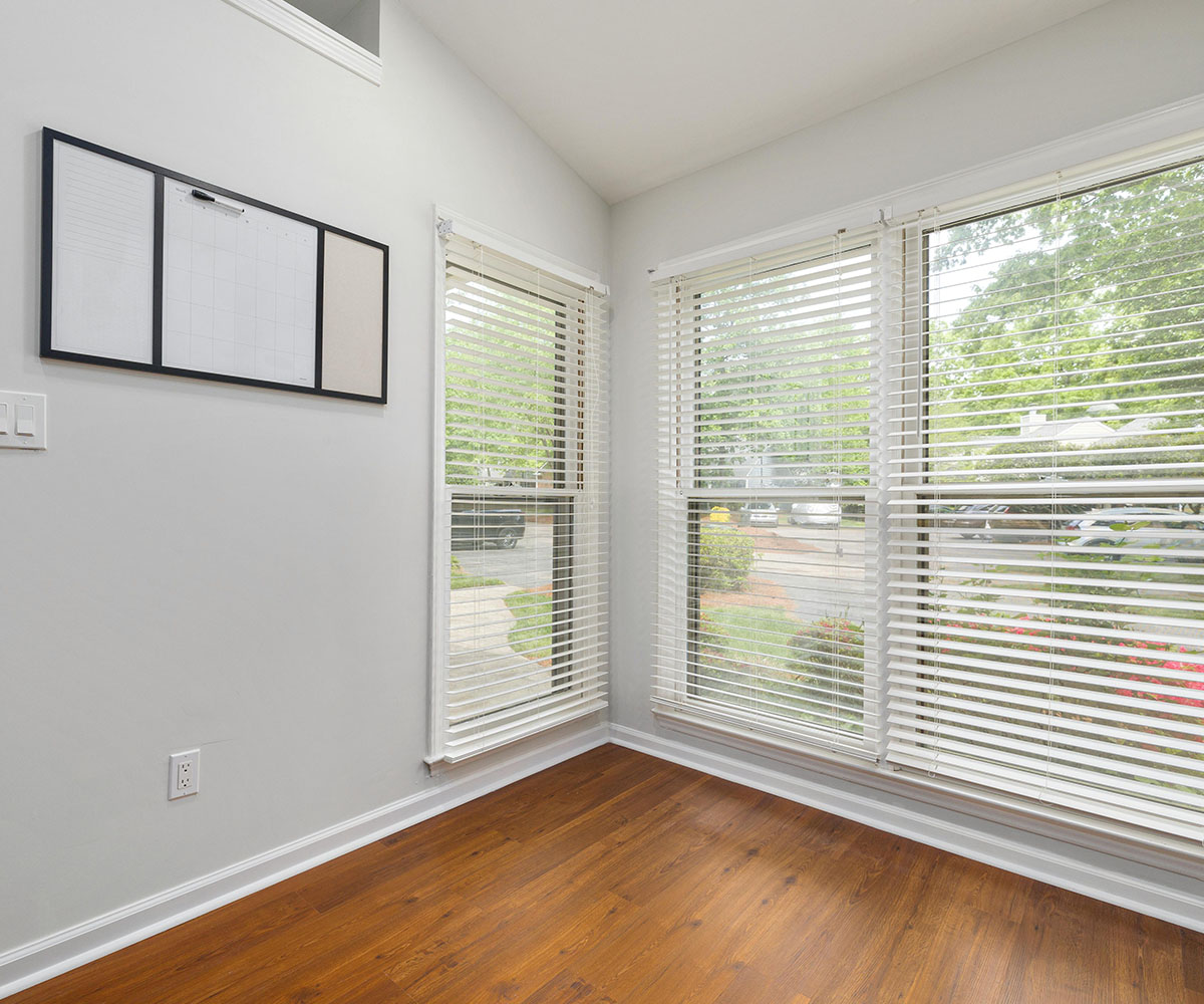 Bright corner office or studio space featuring white horizontal blinds, light grey walls, and warm timber flooring.